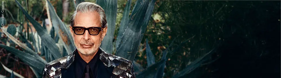 Ein Mann mit Brille und glamourösem Anzug in einer Gartenlandschaft.