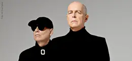 Zwei Personen in schwarzer Kleidung stehen, eine mit Sonnenbrille und Kappe. Beiläufig, neutraler Ausdruck.