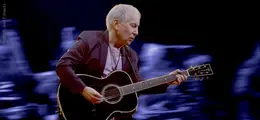 Ein älterer Mann spielt Gitarre vor einem unscharfen, dunklen Hintergrund.
