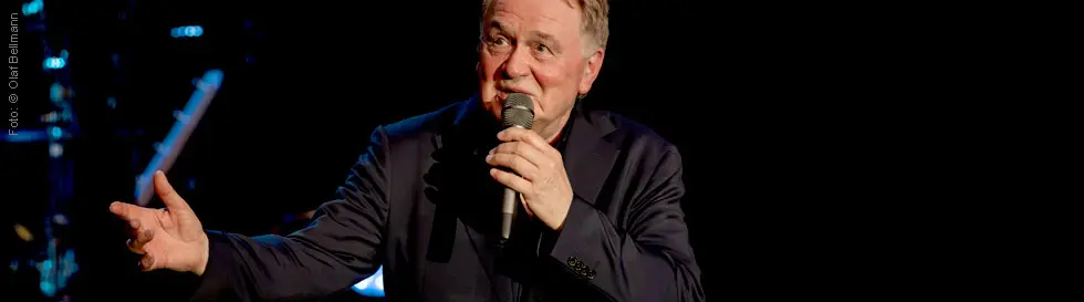 Ein Mann mit Mikrofon singt gestikulierend auf einer Bühne in dunkler Kleidung.