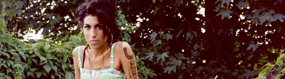 Eine Frau mit dunklem Haar und Tattoos steht vor einem Hintergrund aus dichtem Laub, trägt ein grün-weiß gestreiftes Kleid.