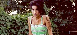 Eine Frau mit dunklem Haar und Tattoos steht vor einem Hintergrund aus dichtem Laub, trägt ein grün-weiß gestreiftes Kleid.