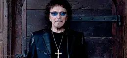 Ein Mann mit dunkler Brille und schwarzer Lederjacke trägt ein großes Kreuz als Kette vor einem Holztor.