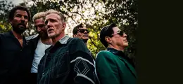 Fünf Männer stehen im Freien vor Bäumen. Sie schauen ernst oder nachdenklich in verschiedene Richtungen.