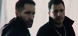 Zwei Männer mit ernstem Blick, dunkle Kleidung, unscharfer Hintergrund.