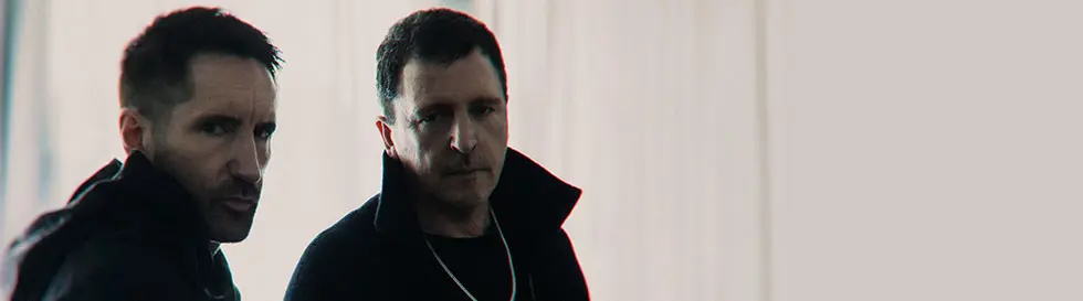 Zwei Männer mit ernstem Blick, dunkle Kleidung, unscharfer Hintergrund.