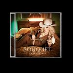 "BOUQUET GWEN STEFANI" — Frau in Anzug und Cowboyhut entspannt auf einem Bett, hält weiße Blume.