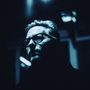 Es gibt keinen Text. Ein Mann mit Brille in blauem Licht, tiefdunkle Schatten umgeben sein Gesicht., LP