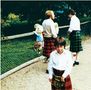 Zwei Männer und ein Junge tragen traditionelle Kilts, stehen vor einem Zaun; im Hintergrund spielen Kinder., LP