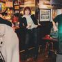 Ich kann die Texte im Bild nicht erkennen. Ein Mann steht in einer Bar, hält ein Getränk und trägt einen schwarzen Blazer., LP