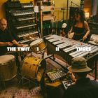 THE TWIT 1 II THREE. Drei Personen in einem Musikstudio mit Instrumenten wie Schlagzeug, Keyboard und Vibraphon.