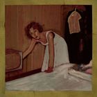 Eine Frau im weißen Kleid beugt sich zu einem alten Fernseher. Ein Kleid hängt an der Wand. Sie wirkt nachdenklich.