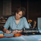Eine Frau in blauer Bluse schreibt konzentriert an einem Tisch, umgeben von Papieren und einer alten Schreibmaschine.