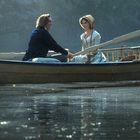 Ein Mann und eine Frau in historischer Kleidung rudern auf einem ruhigen See in einem Boot.