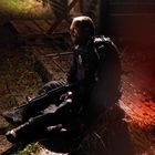 Ein Mann in Uniform sitzt auf nassem Boden, umgeben von schummrigem Licht. Er hält eine Waffe und blickt ernst.