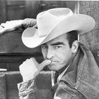 Ein Mann in Cowboyhut und Jeansjacke schaut nachdenklich zur Seite, mit einem Arm auf einem Holzzaun gestützt.
