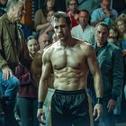 Ein muskulöser Mann in Boxhose steht konzentriert in einer Kampfarena. Umgeben von Zuschauern, die gespannt zusehen.