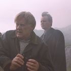 Zwei ältere Männer auf einem nebligen Feld. Der vordere hält ein Fernglas. Beide tragen Mäntel.