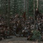 Eine große Gruppe von Menschen in Uniformen steht dicht gedrängt in einem Wald, teils mit Waffen, um eine rote Fahne.