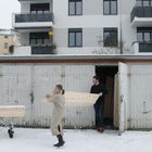 Garagen mit Nr. 1-4. Zwei Personen tragen Holz. Sarg auf Gestell im Schnee, Apartmentgebäude im Hintergrund.