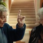 Zwei ältere Männer, einer zeigt mit dem Finger nach oben. Sie stehen vor einer Treppe in einem hellen Gebäude.