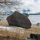 Hinweisschild: Schwimmen verboten. Ein großer Felsen mit Graffiti am sandigen Ufer, gegenüber ein Hafen mit Kränen.