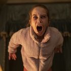 Ein Mädchen in einem flauschigen Pullover schreit mit weit geöffnetem Mund, spitze Zähne sichtbar, in einem dunklen Raum.