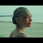 Eine Frau mit Schlamm im Gesicht blickt lächelnd über die Schulter. Hintergrund ist ein ruhiger Strand.