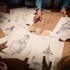 Skizzen von Figuren in historischen Kostümen liegen auf einem Tisch. Mehrere Hände sortieren die Zeichnungen.