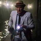 Ein Mann in einem hellen Anzug mit Hut steht in einem Tunnel und hält eine leuchtende Taschenlampe.