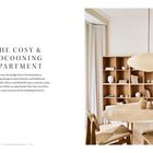 Text: THE COSY & COCOONING APARTMENT. Gemütliches Wohnzimmer mit hellem Holztisch, Stühlen und Regal, schlichte Dekoration.