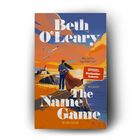 Titel: "The Name Game". Autor: Beth O'Leary. Ein Paar steht an einer Küste, Sonnenuntergang im Hintergrund.