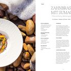 "ZAHNBRASSE MIT SUMACH, Clementinen, Meeresgemüse. Rezept für 4 Personen, Zubereitungszeit: 30 Minuten, Ruhen: 1 Stunde." Links ein Teller mit Gericht auf Steinen am Strand.