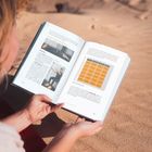 Eine Person liest ein Buch mit Übungen und einer Tabelle, während sie auf einem roten Tuch im Sand sitzt.