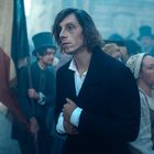 Ein Mann in historischer Kleidung steht ernsthaft inmitten einer Menge, die eine Fahne schwenkt. Eine Frau hebt eine Flasche.