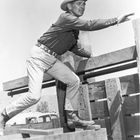 Ein Mann in Cowboy-Kleidung klettert über Holzplanken. Er trägt einen Hut, Stiefel und einen Gürtel mit Verzierungen.