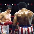Zwei Boxer stehen sich im Ring gegenüber, der eine mit roten Handschuhen und weiß-roter Hose, der andere in Blau-Roten.
