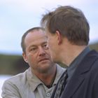 Zwei Männer im Gespräch; der eine ist älter mit Bart und trägt einen hellen Mantel, der andere einen dunklen Anzug. Hintergrund: Natur.