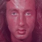 Es gibt keinen sichtbaren Text. Ein Mann mit langen Haaren und ernstem Gesichtsausdruck ist zu sehen, rötliche Töne dominieren.