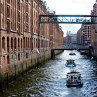 Auf einem Kanal in Hamburg fahren Boote zwischen hohen Backsteingebäuden. Über dem Wasser sind Brücken.