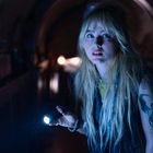 Keine sichtbaren Texte. Eine blonde Frau mit Taschenlampe in einem dunklen Raum. Sie hat ein Tattoo am Arm.