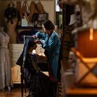 Eine Frau im blauen Kittel arbeitet an einer sitzenden Person in einem Schneideratelier mit Schneiderpuppen und Stoffen.