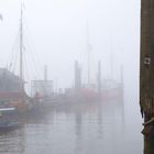 „GUSTAV VOGEL“ steht auf einem Schiffsrumpf. Dunstiger Hafen, Schiffe teils verdeckt, Holzmast im Vordergrund mit Tau.