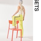 Der Text "SETS" steht am rechten Rand. Eine Person mit grauem Haar steht barfuß auf einem orangefarbenen Stuhl, trägt ein helles Top und gelbe Hosen.