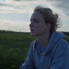 Eine Person mit einem grauen Kapuzenpullover schaut nachdenklich zur Seite, im Hintergrund ist ein unscharfes Feld und bewölkter Himmel.