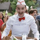 Ein Mann in Retro-Eismacher-Uniform mit roter Fliege und Papphut verteilt Getränke an fröhliche Kinder.