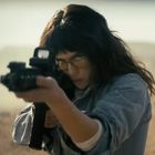Keine Texte. Eine Person mit Brille zielt mit einem Gewehr in einer weiten, trockenen Landschaft.