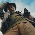 Ein älterer Mann mit Hut und Bart hält ein Huhn, schaut ernst in einem abgenutzten Outfit nach unten. Himmel im Hintergrund.