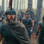 Ein Mann in antiker Rüstung mit Helm führt eine Gruppe Krieger durch einen Wald. Sie tragen Schwerter und Schilde.