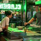 Neon-Schriftzug: "CLAREMONT". Eine Frau lehnt sich lächelnd zu einem Mann, der eine Pizza zubereitet. Warmes, grünes Licht.